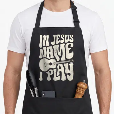 Play acoustic christian apron - Retro cream lettering reading IN JESUS NAME I PLAY with a horizontal acoustic guitar — shown on a black cotton canvas apron against a white background