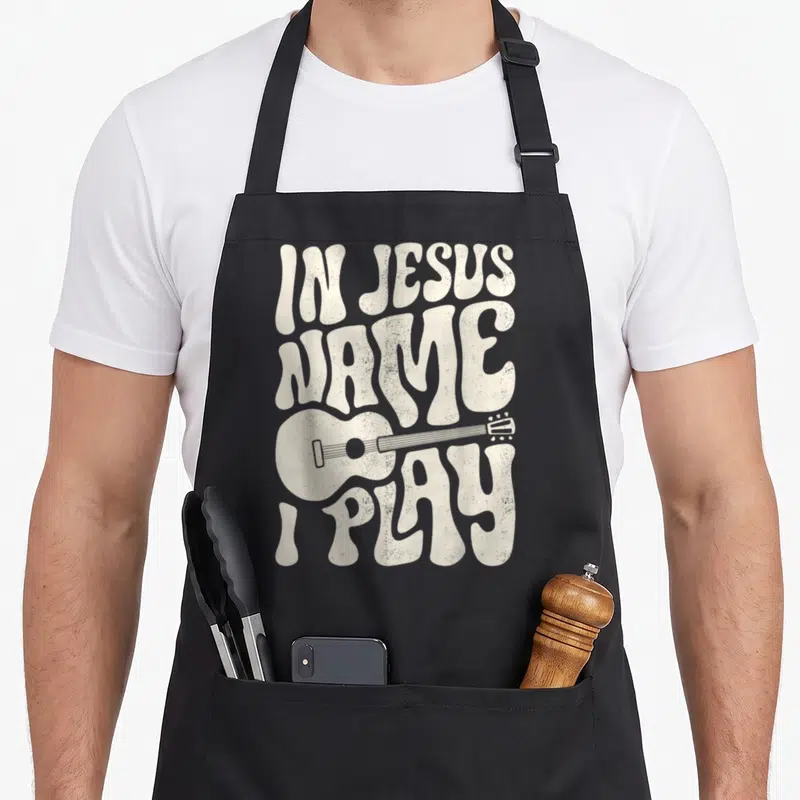 Play acoustic christian apron - Retro cream lettering reading IN JESUS NAME I PLAY with a horizontal acoustic guitar — shown on a black cotton canvas apron against a white background