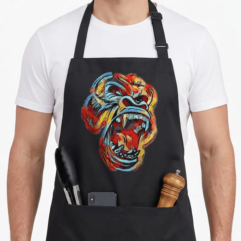 Colourful ape apron - Roaring gorilla head in red orange yellow and blue cartoon style — shown on a black cotton canvas apron against a white background