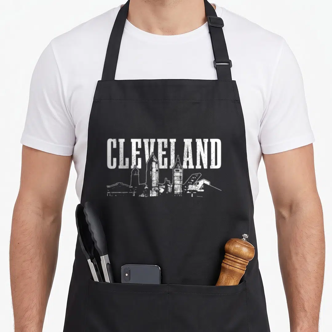 Ohio skyline pride apron - White distressed CLEVELAND text over city skyline silhouette — shown on a black cotton canvas apron against a white background