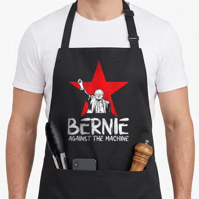 Bernie machine apron - Red star with white Bernie Sanders illustration and distressed BERNIE AGAINST THE MACHINE text — shown on a black cotton canvas apron against a white background