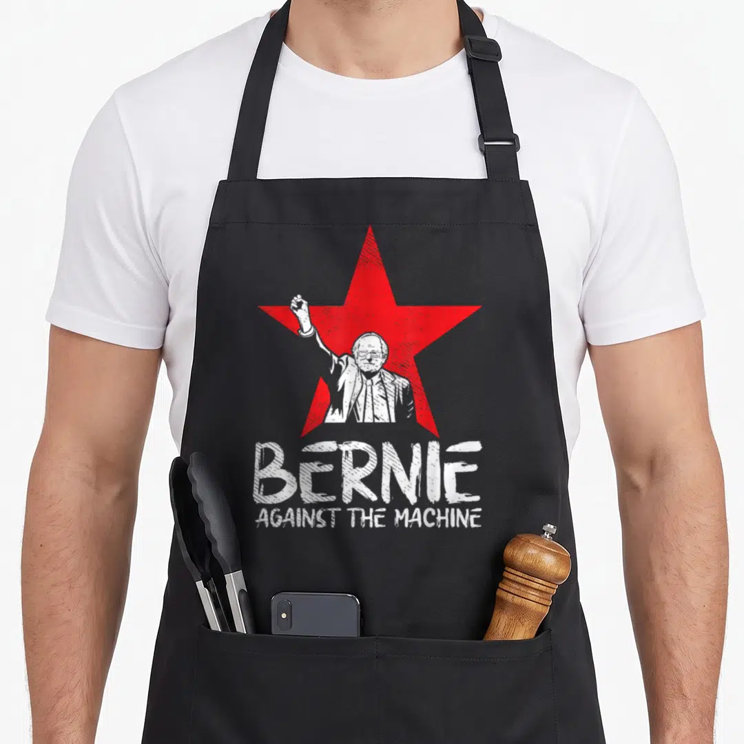 Bernie machine apron - Red star with white Bernie Sanders illustration and distressed BERNIE AGAINST THE MACHINE text — shown on a black cotton canvas apron against a white background
