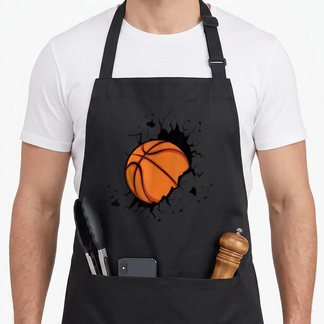 Basketball sports team aprons - Orange basketball bursting through black cracked wall in cartoon style — shown on a black cotton canvas apron against a white background