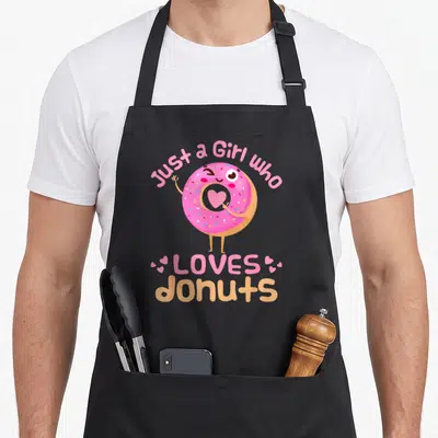 Cute doughnut donut apron - Pink cartoon donut with blue sprinkles pointing to a heart in its hole surrounded by text — shown on a black cotton canvas apron against a white background