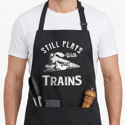 Playing locomotive railroad apron - White steam locomotive with arched text Still Plays With Trains in retro style — shown on a black cotton canvas apron against a white background