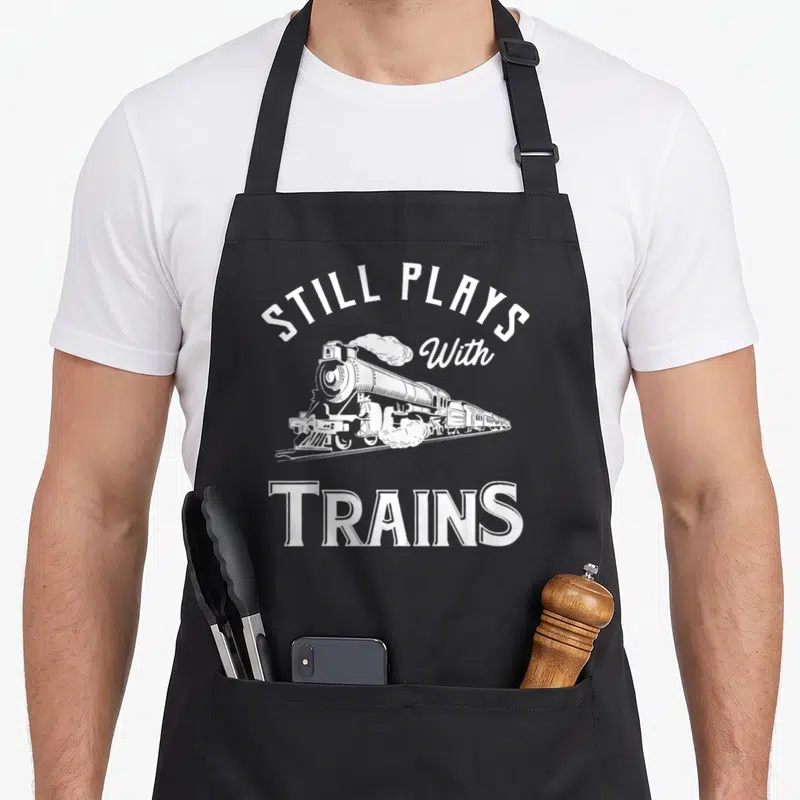 Playing locomotive railroad apron - White steam locomotive with arched text Still Plays With Trains in retro style — shown on a black cotton canvas apron against a white background