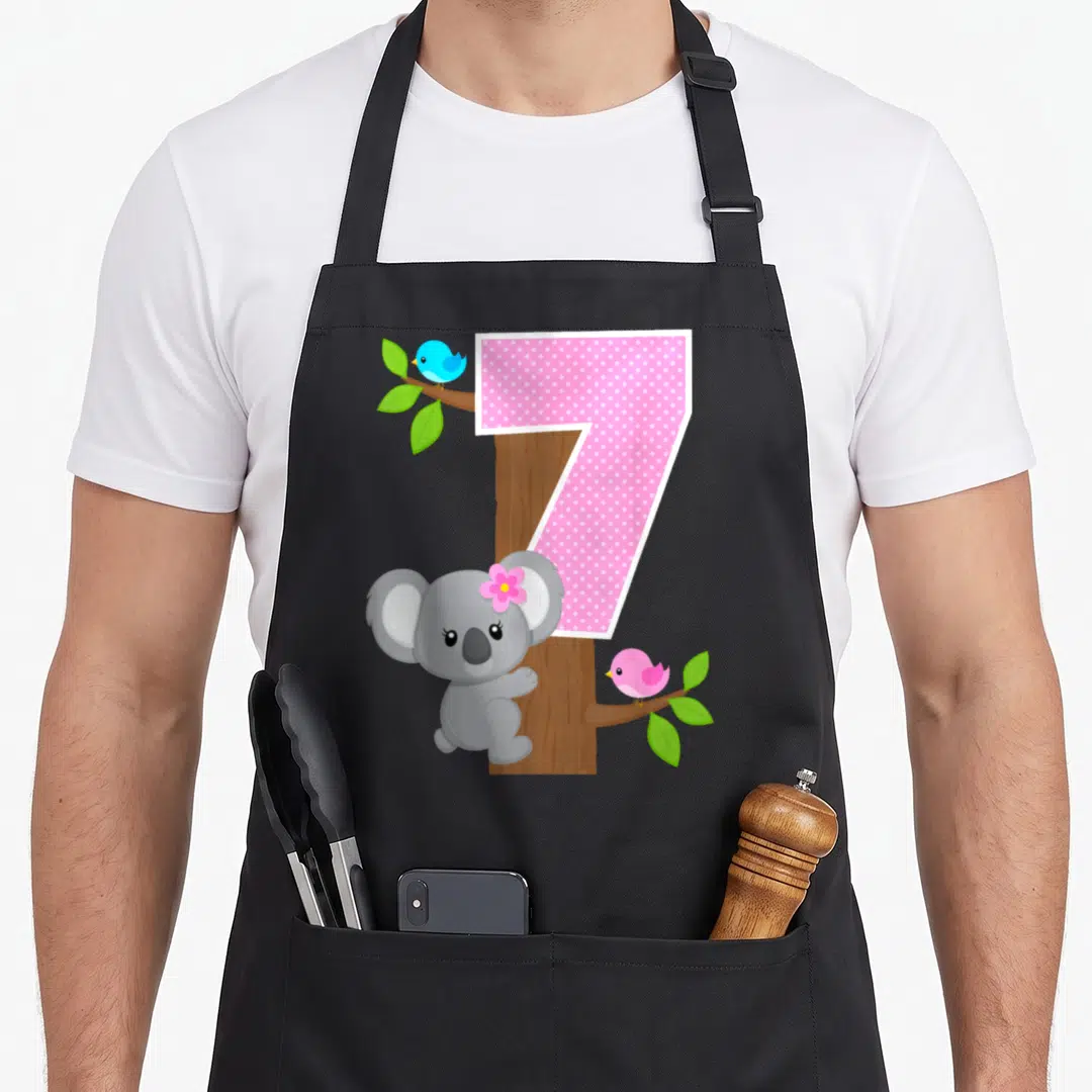 7th bear apron - Pink polka dot number seven over brown tree trunk with grey koala and blue and pink birds — shown on a black cotton canvas apron against a white background