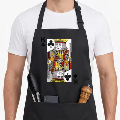 Blackjack card king apron - Mirrored King of Clubs playing card print with K and club symbols in red yellow white — shown on a black cotton canvas apron against a white background