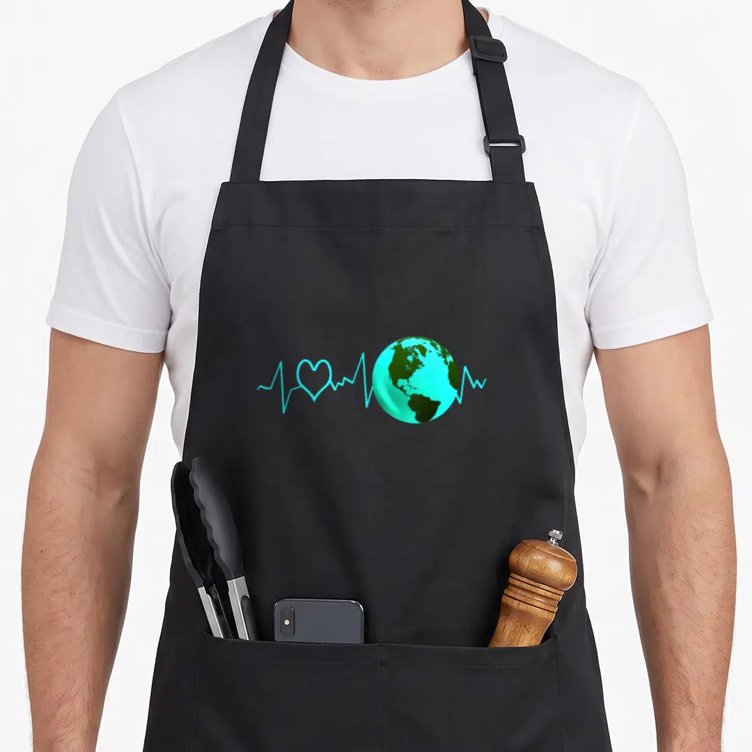 Heartbeat change apron - Cartoon Earth with cyan heartbeat line and heart icon on front panel — shown on a black cotton canvas apron against a white background