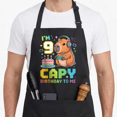 9th bday birthday apron - Cartoon brown capybara with rainbow headphones and green controller next to cake and I'M 9 — shown on a black cotton canvas apron against a white background