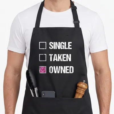 Bdsm owner apron - Checklist with SINGLE TAKEN OWNED text and pink box with white heart — shown on a black cotton canvas apron against a white background