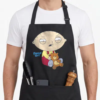 Stewie iconic aprons - Stewie Griffin sitting holding brown teddy bear with Family Guy logo in yellow red blue and brown — shown on a black cotton canvas apron against a white background