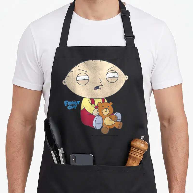 Stewie iconic aprons - Stewie Griffin sitting holding brown teddy bear with Family Guy logo in yellow red blue and brown — shown on a black cotton canvas apron against a white background
