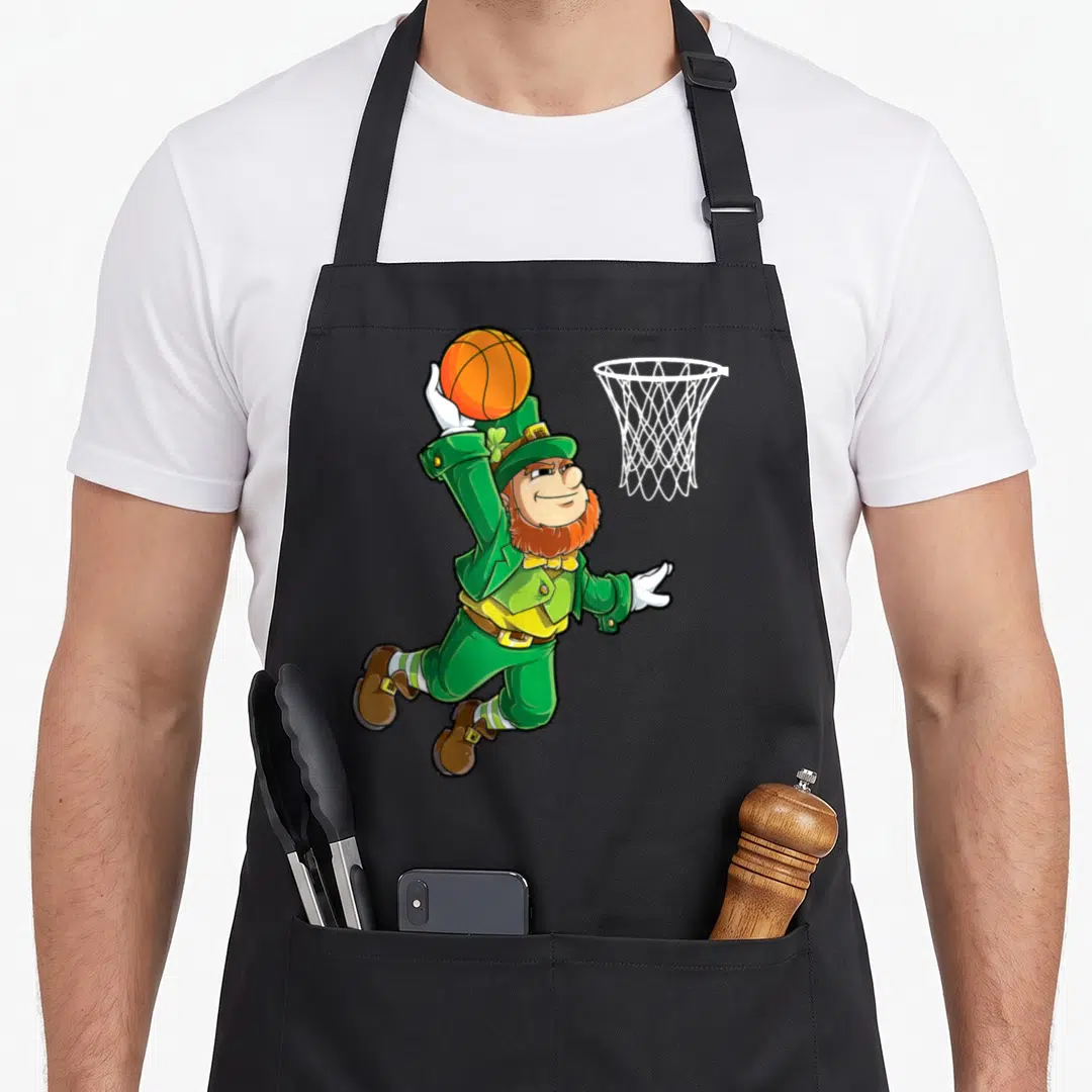 Basketball st patrick day aprons - Cartoon leprechaun jumping to dunk orange basketball into white hoop — shown on a black cotton canvas apron against a white background
