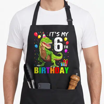 Rex birthday apron - Green T-Rex wearing party hat with IT'S MY 6 TH BIRTHDAY text and colorful balloons — shown on a black cotton canvas apron against a white background