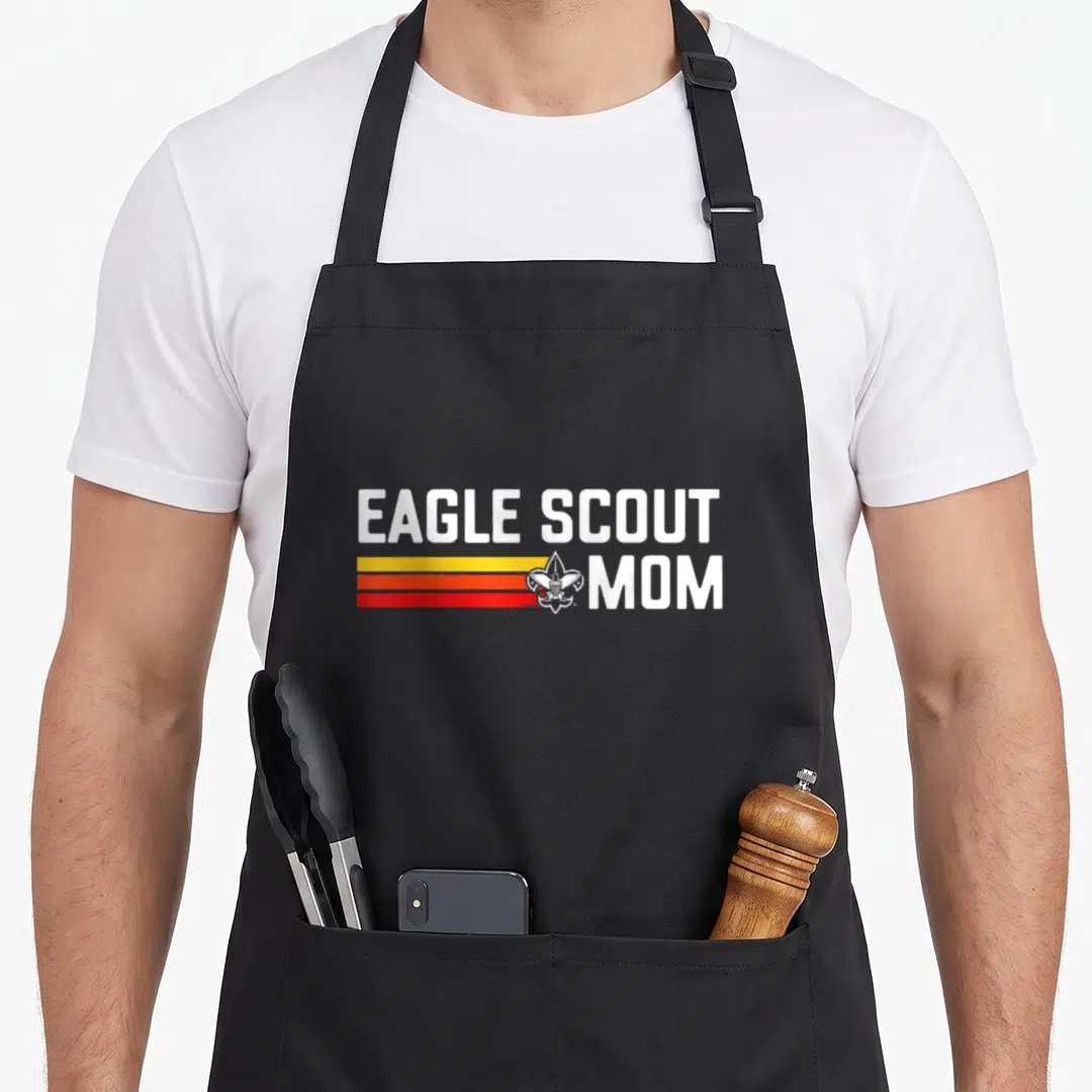Scout pride 80s eagles apron - EAGLE SCOUT MOM text with yellow orange red stripes and emblem overlapping — shown on a black cotton canvas apron against a white background