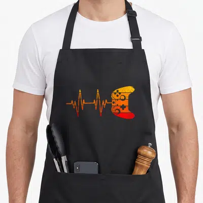 Gamer heartbeat gaming video game apron - Orange and red EKG line transitioning into a game controller silhouette — shown on a black cotton canvas apron against a white background