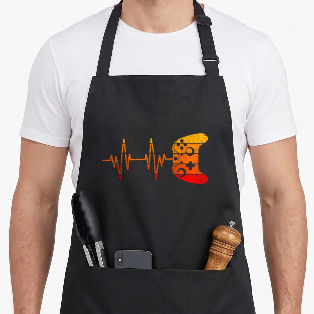 Gamer heartbeat gaming video game apron - Orange and red EKG line transitioning into a game controller silhouette — shown on a black cotton canvas apron against a white background