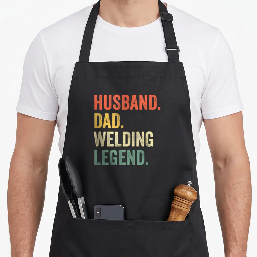 Welder husband father day apron - Stacked red yellow beige and green distressed text reading Husband Dad Welding Legend — shown on a black cotton canvas apron against a white background