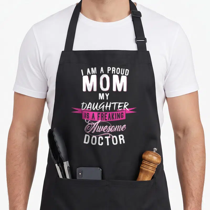 Proud doctor mom daughter apron - Stacked white text reading I am a proud mom my daughter is a freaking awesome doctor — shown on a black cotton canvas apron against a white background