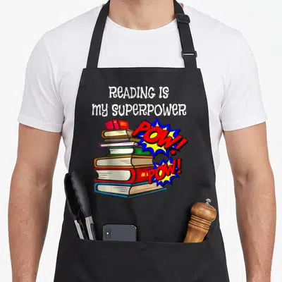 Reading english apron - Stack of colorful books with two POW! bursts and white text reading READING IS my SUPERPOWER — shown on a black cotton canvas apron against a white background