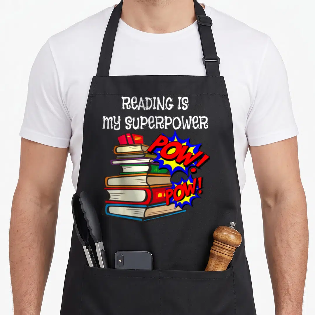 Reading english apron - Stack of colorful books with two POW! bursts and white text reading READING IS my SUPERPOWER — shown on a black cotton canvas apron against a white background