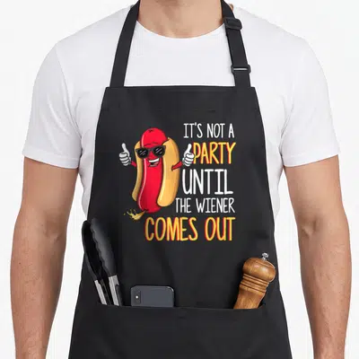 Wiener hot dog apron - Cartoon hot dog with sunglasses thumbs up and fire, stacked text IT'S NOT A PARTY UNTIL — shown on a black cotton canvas apron against a white background