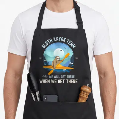 Kayak sloth apron - Cartoon sloth in orange kayak with text SLOTH KAYAK TEAM EST 1983 Relax We Will Get There — shown on a black cotton canvas apron against a white background