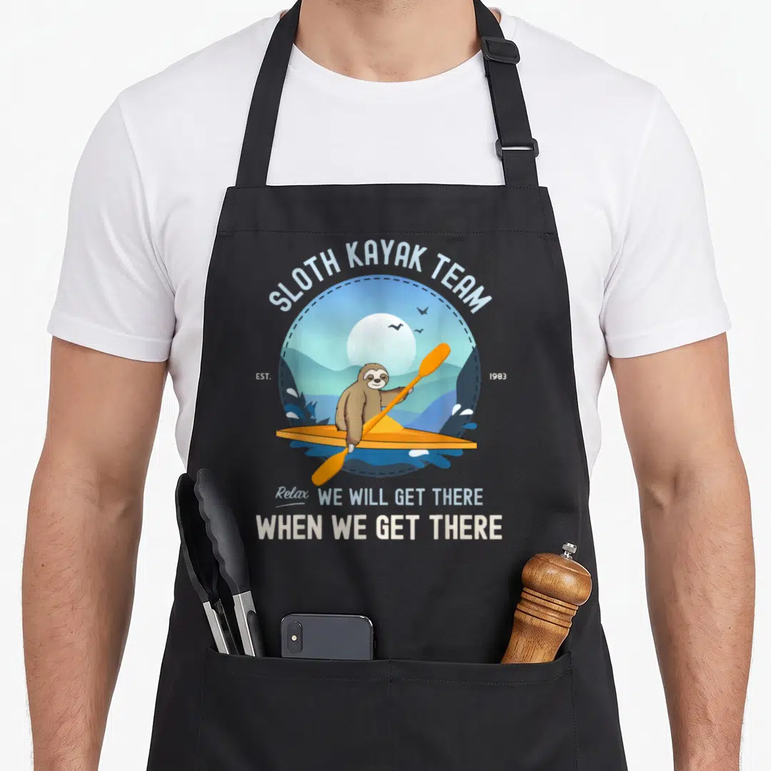 Kayak sloth apron - Cartoon sloth in orange kayak with text SLOTH KAYAK TEAM EST 1983 Relax We Will Get There — shown on a black cotton canvas apron against a white background
