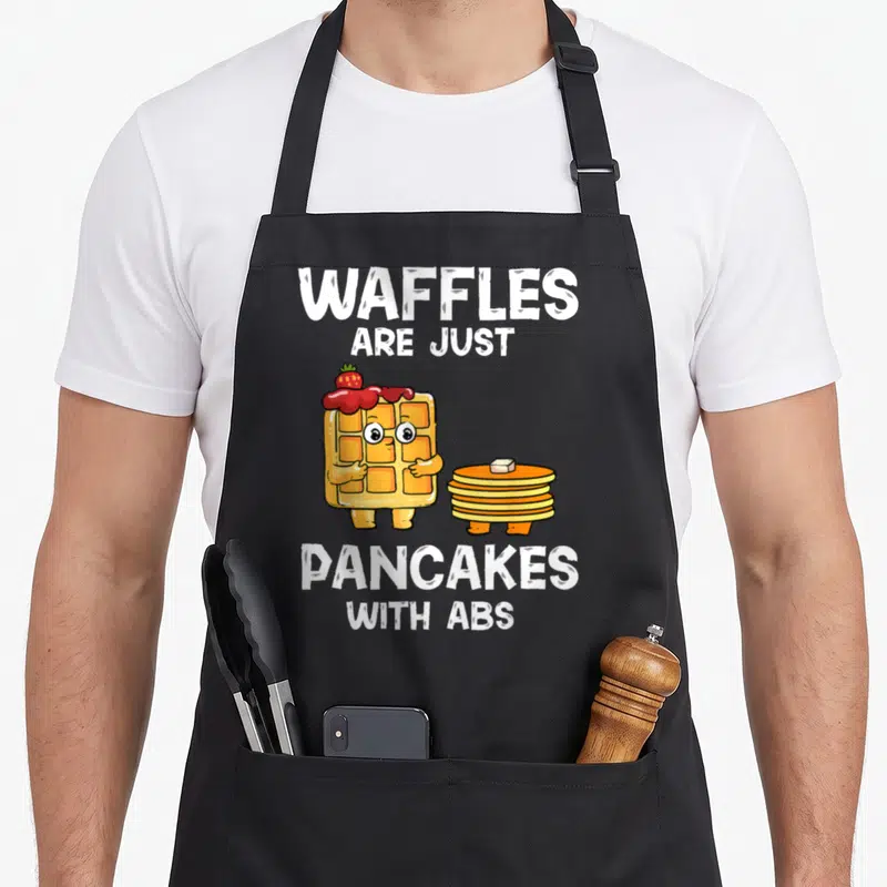 Abs pancake apron - Cartoon waffle with strawberry syrup standing beside a stack of pancakes with butter and white text — shown on a black cotton canvas apron against a white background