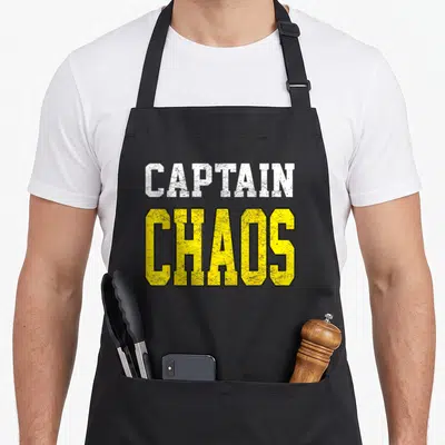 Captain teacher apron - White and yellow distressed block letters reading CAPTAIN CHAOS stacked vertically — shown on a black cotton canvas apron against a white background