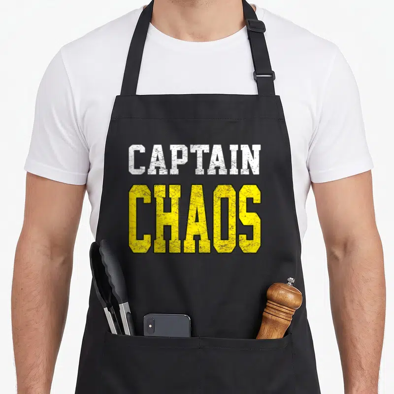 Captain teacher apron - White and yellow distressed block letters reading CAPTAIN CHAOS stacked vertically — shown on a black cotton canvas apron against a white background