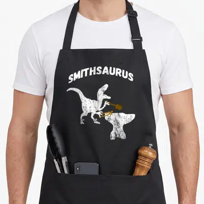 Sledge blacksmith apron - White Smithsaurus text above standing dinosaur holding gold hammer and tongs over anvil — shown on a black cotton canvas apron against a white background