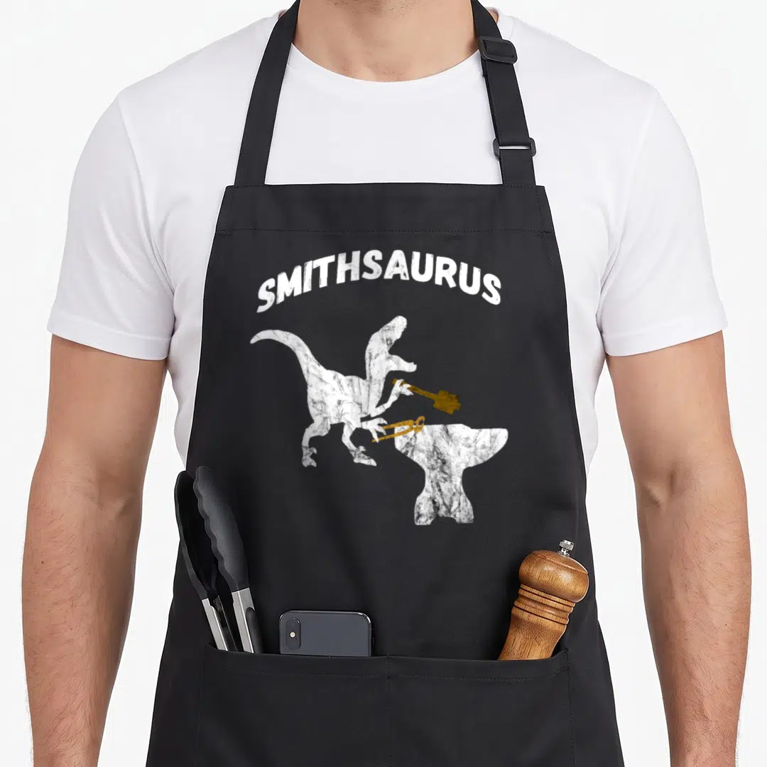 Sledge blacksmith apron - White Smithsaurus text above standing dinosaur holding gold hammer and tongs over anvil — shown on a black cotton canvas apron against a white background