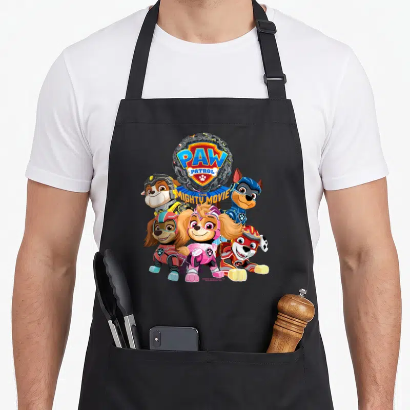 Mighty patrol happy apron - PAW PATROL THE MIGHTY MOVIE logo above four cartoon puppies in colorful superhero suits — shown on a black cotton canvas apron against a white background