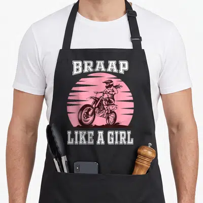 Braap bike apron - Motocross rider on bike under pink sun with BRAAP LIKE A GIRL text in white pink brown — shown on a black cotton canvas apron against a white background