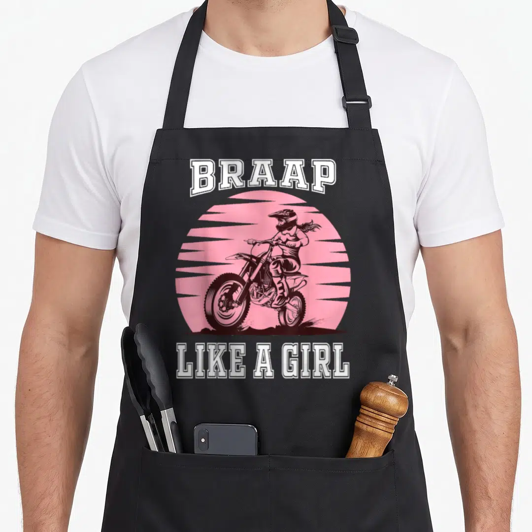 Braap bike apron - Motocross rider on bike under pink sun with BRAAP LIKE A GIRL text in white pink brown — shown on a black cotton canvas apron against a white background