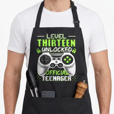 13th birthday video game apron - Lime green controller with LEVEL THIRTEEN UNLOCKED OFFICIAL TEENAGER text and plus — shown on a black cotton canvas apron against a white background