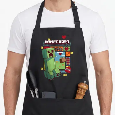 Creeper pixel apron - MINECRAFT logo, green Creeper, red TNT, hearts, and blocks in pixel art style — shown on a black cotton canvas apron against a white background