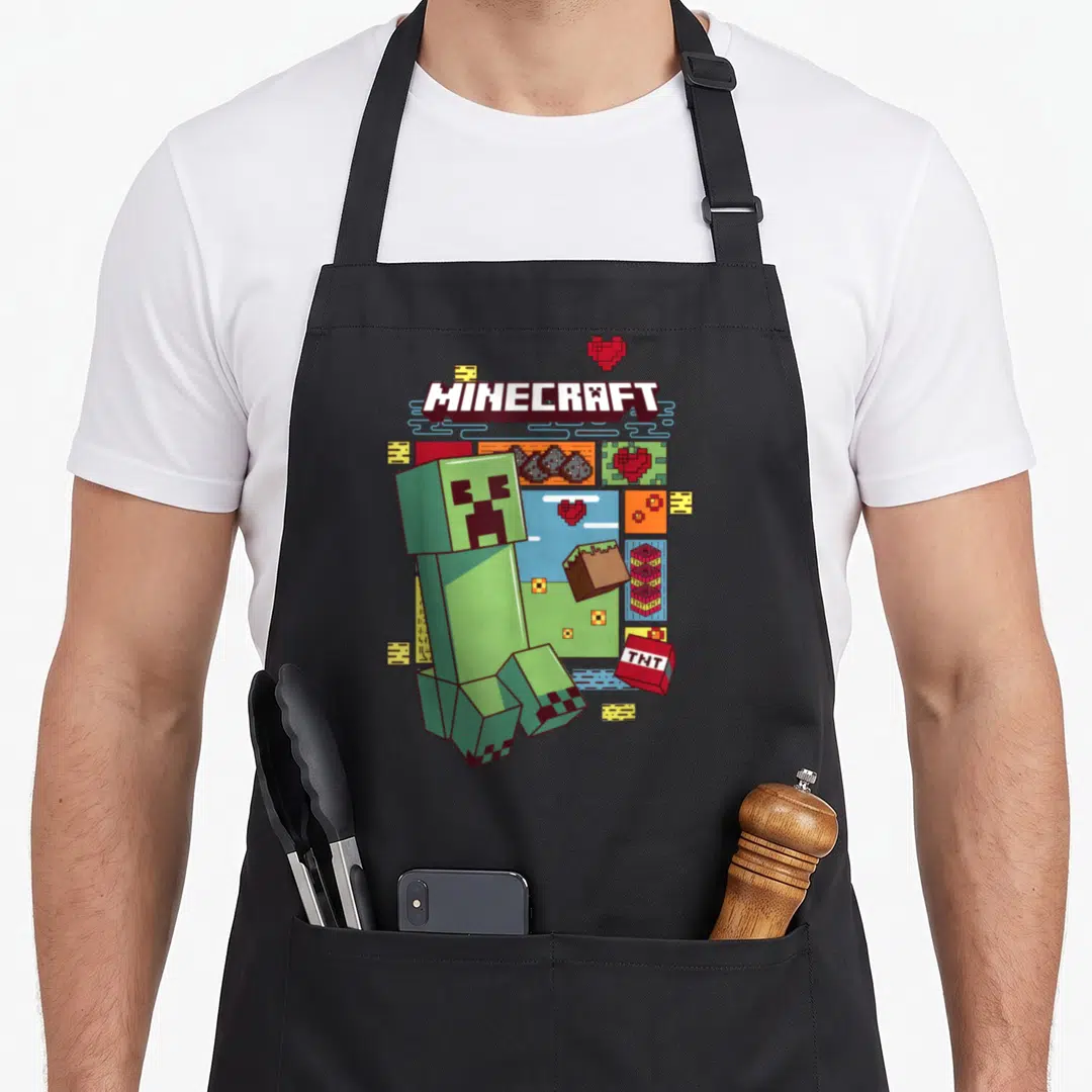 Creeper pixel apron - MINECRAFT logo, green Creeper, red TNT, hearts, and blocks in pixel art style — shown on a black cotton canvas apron against a white background
