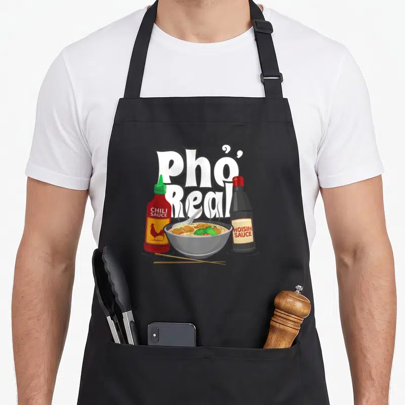 Bowl pho apron - Cartoon pho bowl with chili and hoisin sauce bottles and Pho Real text — shown on a black cotton canvas apron against a white background