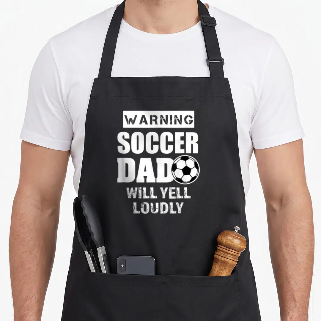 Yell soccer apron - Stacked warning text with soccer ball overlapping the word dad in distressed white print — shown on a black cotton canvas apron against a white background