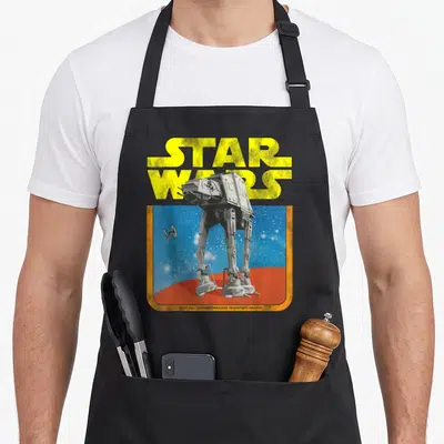 Walker tie apron - Yellow STAR WARS text above grey AT-AT walker on red ground with TIE fighter in blue starry sky — shown on a black cotton canvas apron against a white background