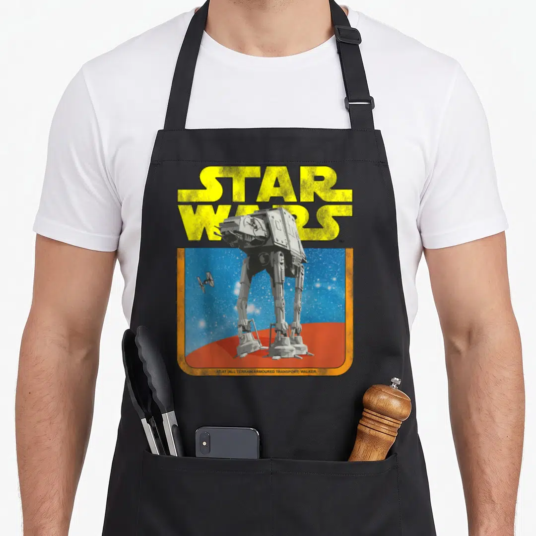 Walker tie apron - Yellow STAR WARS text above grey AT-AT walker on red ground with TIE fighter in blue starry sky — shown on a black cotton canvas apron against a white background