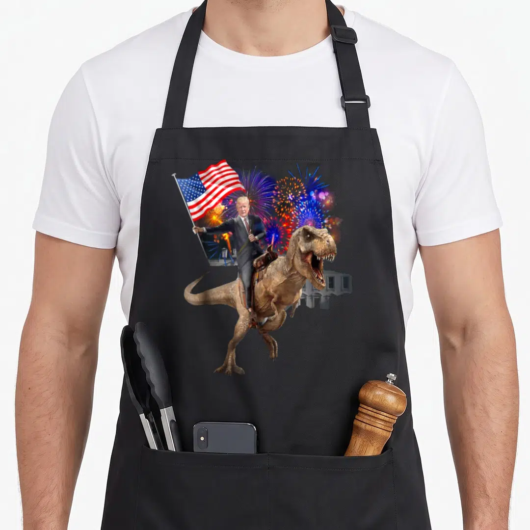 Trump dinosaur apron - Donald Trump riding a T-Rex holding a USA flag with fireworks and White House in red white blue — shown on a black cotton canvas apron against a white background