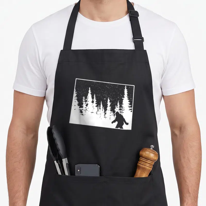 Outdoor bigfoot apron - Grey Bigfoot silhouette in snowy pine forest, minimalist design — shown on a black cotton canvas apron against a white background