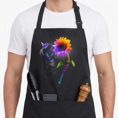 Rheumatoid sunflower apron - Rainbow sunflower with hummingbird, scattered teal ribbons, and curved awareness text — shown on a black cotton canvas apron against a white background