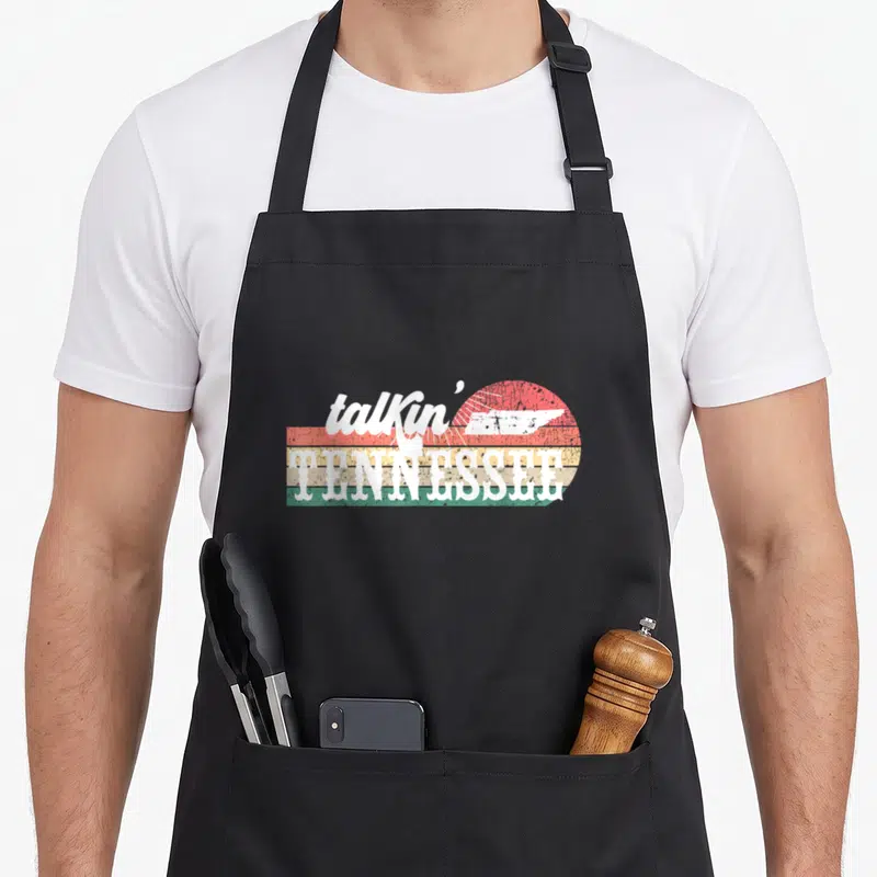 Talkin country kitchen apron - White talkin Tennessee text over red orange yellow green striped sun with state — shown on a black cotton canvas apron against a white background