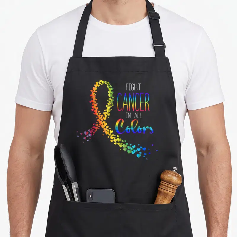 Suck cancer man apron - Rainbow ribbon made of hearts next to stacked text FIGHT CANCER IN ALL COLORS — shown on a black cotton canvas apron against a white background
