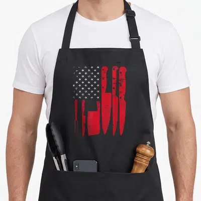 Butcher american flag apron - Red knives and cleavers arranged as a US flag with white stars on grey background — shown on a black cotton canvas apron against a white background